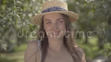 可爱的时尚女青年戴着草帽，穿着白色连衣裙，看着站在绿色夏日花园里的相机